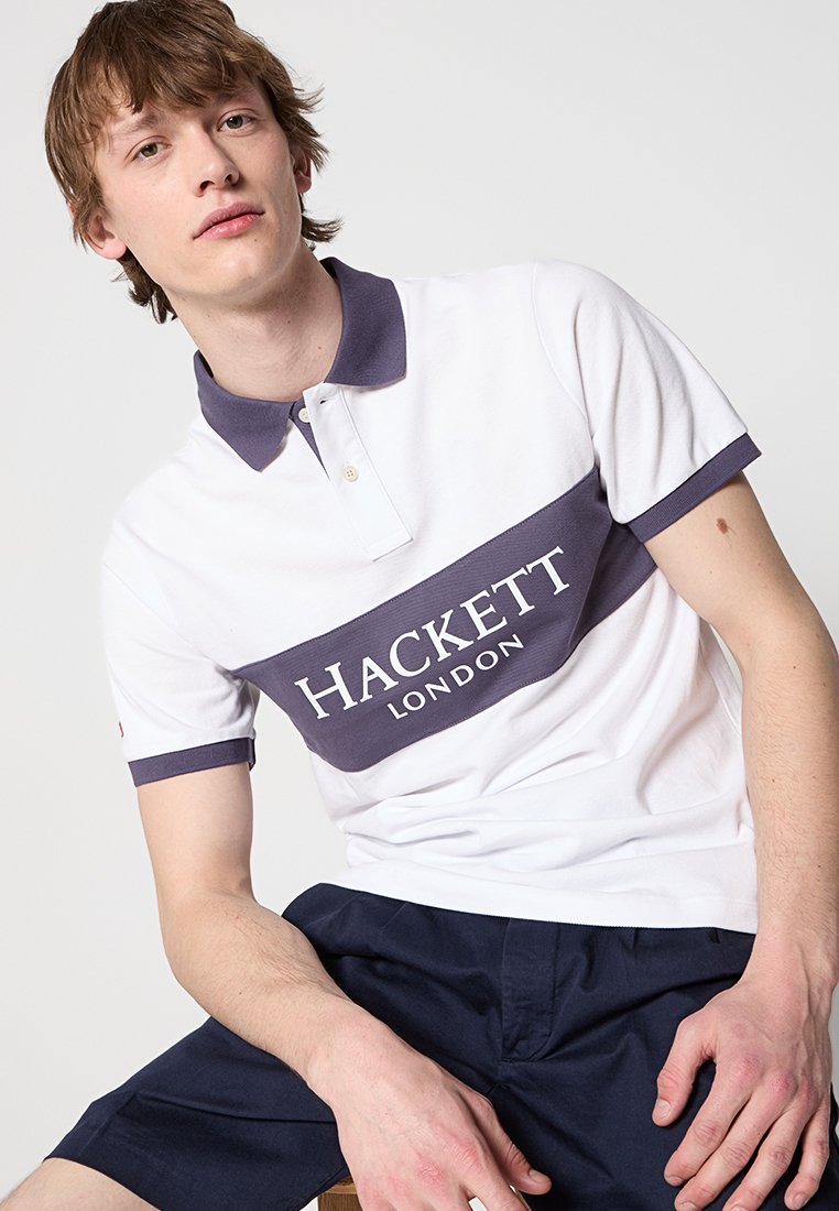 Polo shirt avec un corps blanc et un col et un ourlet bleu marine. Présente une rayure horizontale bleu marine avec le texte "HACKETT LONDON" en blanc.