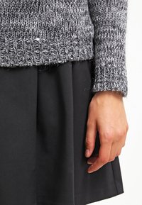 Mano appoggiata accanto a una gonna nera, indossando un maglione lavorato a maglia grigio e bianco con polsini e fondo a coste.