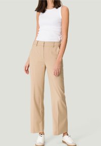 Pantalon beige à fines rayures avec une coupe ajustée, doté de passants pour ceinture et d'une fermeture éclair, associé à un débardeur blanc et des baskets blanches.