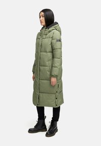 Lang, polstret grøn pufferjakke med hætte, der har en sort foring. Den har en lige pasform, frontknaplukning og er parret med sorte støvler.