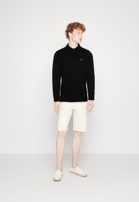 Black long-sleeve polo shirt, tailored fit, subtle logo on chest, paired with cream-colored shorts and light slip-on shoes.