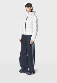 Hvid pufferjakke med lynlås foran og hætte; parret med brede mørkeblå denimbukser med sideliner og frynset kant.