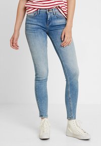 Jeans ajustados en denim azul claro, con un diseño clásico de cinco bolsillos y una cremallera. Combinados con zapatillas deportivas blancas para crear contraste.