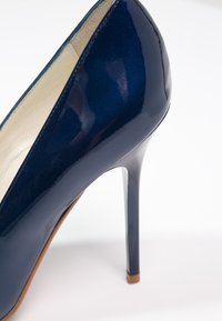 Chaussure à talon haut en cuir verni bleu foncé, avec un bout pointu et un talon aiguille élancé, sur fond blanc.