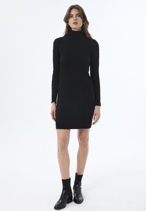 Robe noire tricotée avec un col montant, présentant un motif en maille torsadée et des manches longues, assortie à des bottines noires à talons.