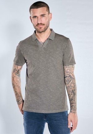 Mann mit kurzen Haaren und vollarmigen Tattoos, der ein graues kurzärmeliges Poloshirt und blaue Jeans trägt, steht vor hellem Hintergrund.