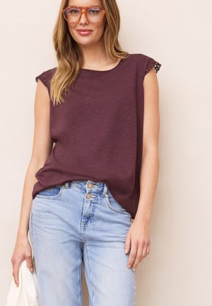 Kvinde med langt blondt hår iført en ærmeløs vinrød top, lyseblå højtaljede jeans, orange briller og holder et hvidt stykke stof.