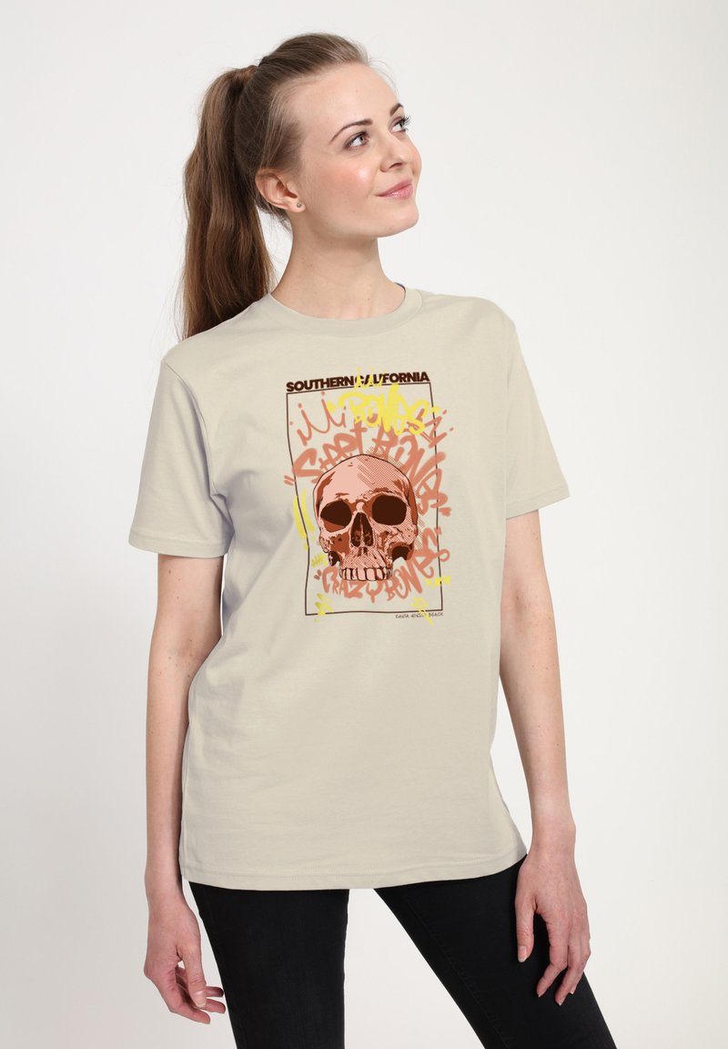 Beige katoenen t-shirt met een centrale doodskopgrafiek en graffiti-stijl tekst in oranje en gele accenten. Korte mouwen en een ontspannen pasvorm.