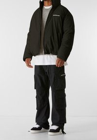 Giacca a vento nera con colletto alto, maglione grigio sotto, pantaloni cargo neri e sneakers nere con lacci bianchi.