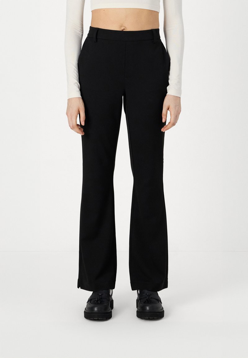 Pantalons évasés noirs à coupe ajustée, dotés de poches avant, de passants de ceinture et d'une texture de tissu lisse. Portés avec des chaussures noires.