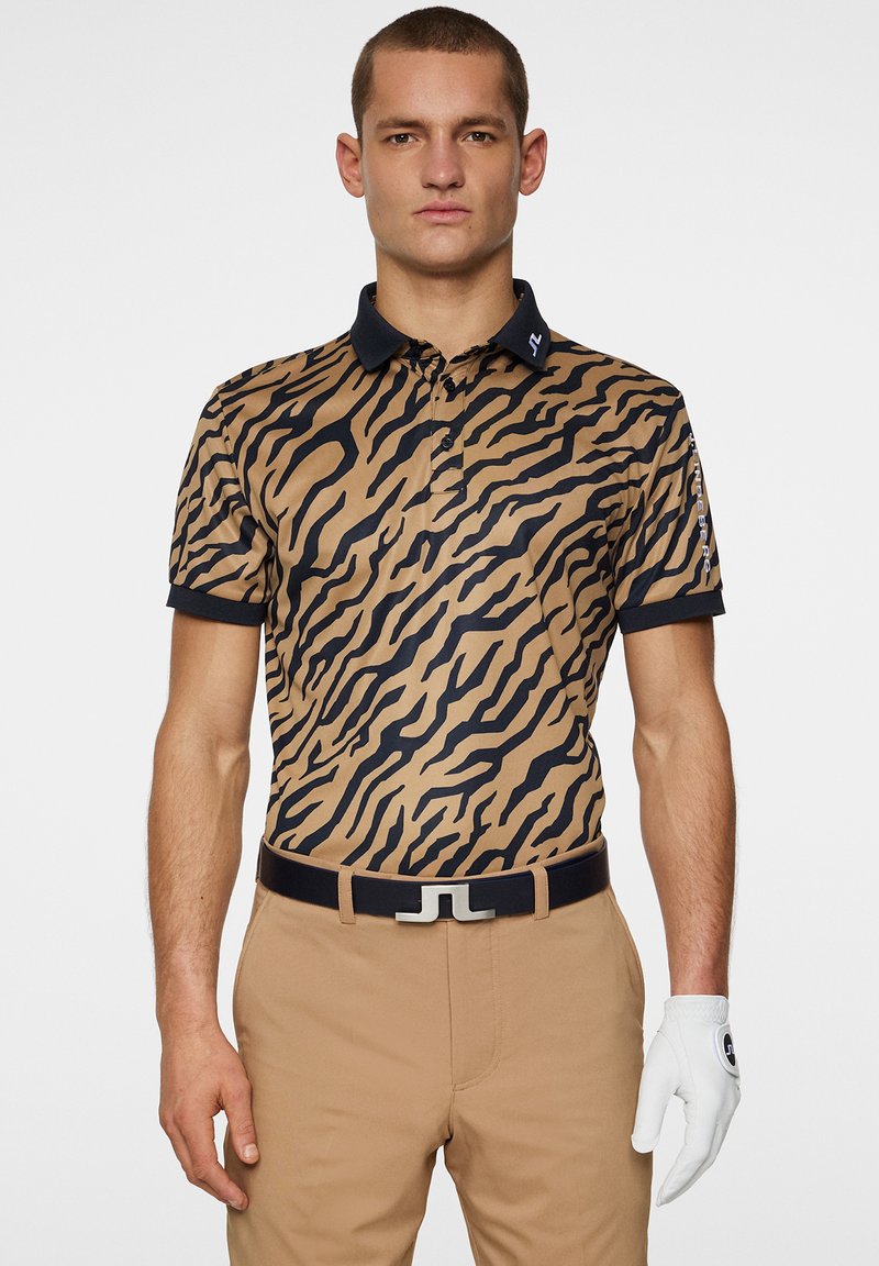 Polohemd in Tan mit schwarzen Tigerstreifen, kurzen Ärmeln und einem marineblauen Kragen. Kombiniert mit beigen Hosen und einem schwarzen Gürtel mit einer silbernen Schnalle.