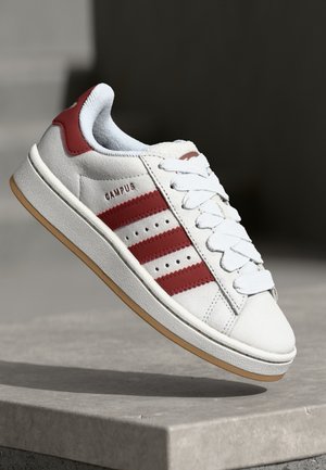 Biały sneaker z czerwonymi paskami i detalami, białe sznurowadła, napis "CAMPUS" po boku, beżowa podeszwa, przedstawiony na kamiennej powierzchni na ciemnym tle.