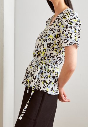 Kvinde iført en kortærmet, mønstret bluse med bindebælte i taljen, kombineret med sorte bukser, foran en ensfarvet lys baggrund.