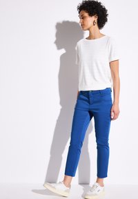 Blaue, enganliegende Hosen mit glatter Textur, fünf Taschen und einem Knopfverschluss, kombiniert mit einem lässigen weißen Oberteil mit Struktur und hellen Sneakers.