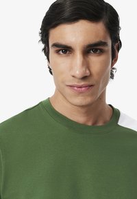 Felpa verde in cotone con colletto girocollo, texture liscia e accento bianco sulla spalla. Inquadratura ravvicinata della parte superiore del torso di un modello maschile.