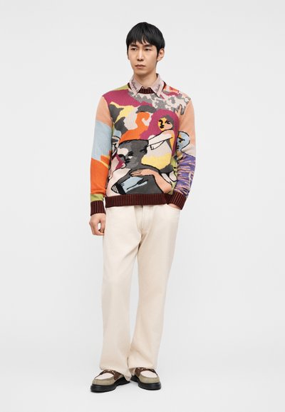 Πλεκτό πουλόβερ με πολύχρωμα αφηρημένα σχέδια σε πολλαπλές αποχρώσεις, σε συνδυασμό με μπεζ παντελόνια και casual παπούτσια, αναδεικνύοντας μοναδική καλλιτεχνική έκφραση.