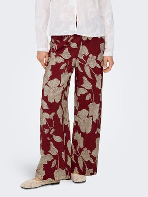 Vrouw draagt wijdere bordeauxrode broek met beige bloemenprint, gecombineerd met een witte blouse en beige instapschoenen.