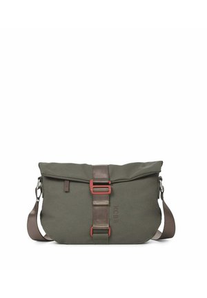 Femme aux cheveux blonds attachés en chignon, portant un pull marron et un pantalon, avec un sac bandoulière gris à sangle beige, marchant vers la gauche.
