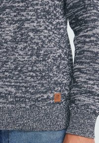 Acercamiento a la manga y el dobladillo de un suéter de punto en blanco y negro con una pequeña etiqueta de cuero marrón de la marca "Blend", usado con jeans azules.