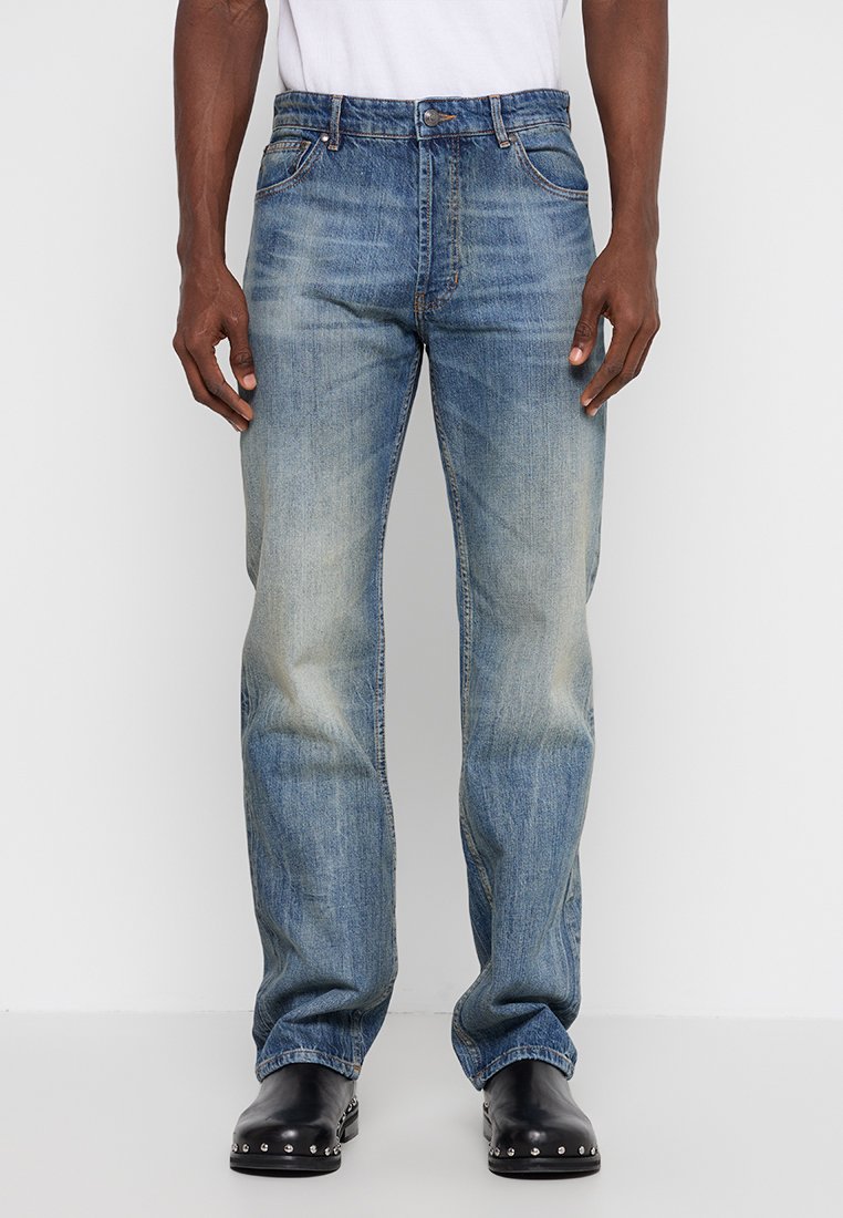 Homme portant un jean droit bleu délavé et des chaussures en cuir noires à clous, se tenant devant un fond blanc uni.