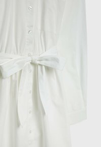 Robe blanche en tissu léger avec un devant à boutons, des manches longues et une taille ceintrée. Dispose d'une poche poitrine et d'une texture lisse.