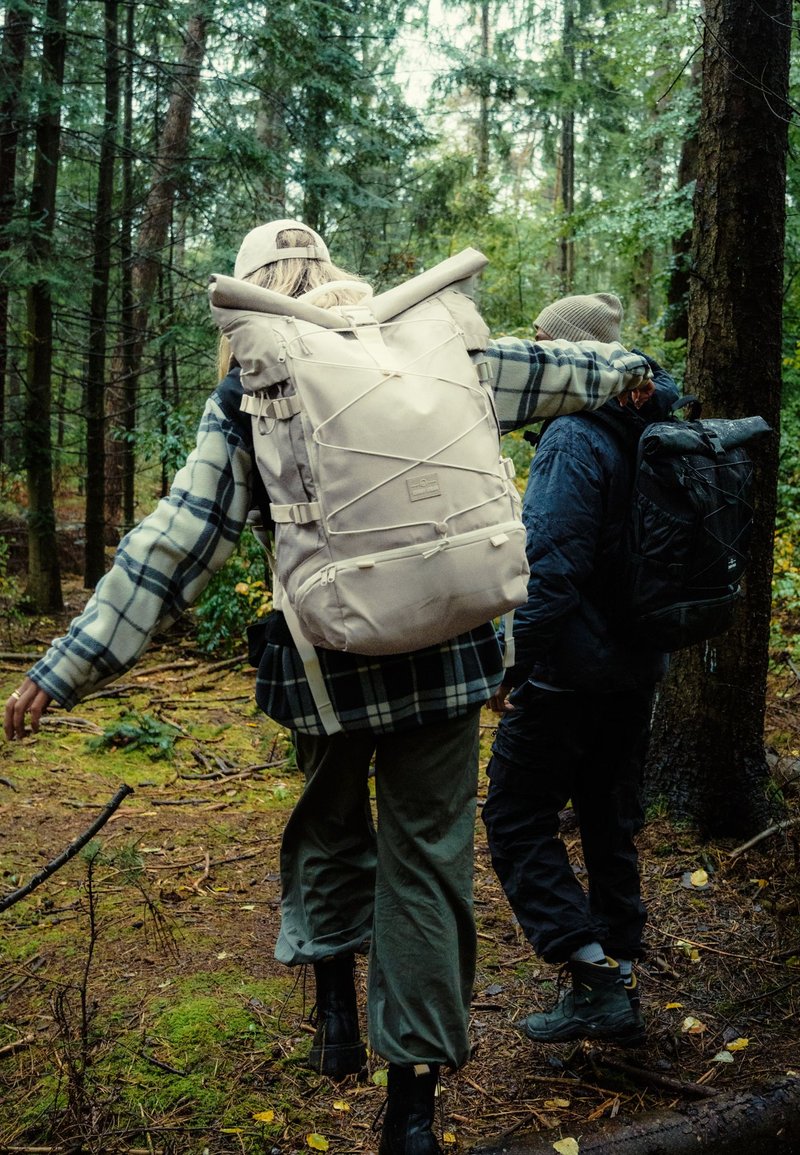 Két túrázó, nagy hátizsákokkal, sűrű, zöld erdőben sétál egy moha borította ösvényen.