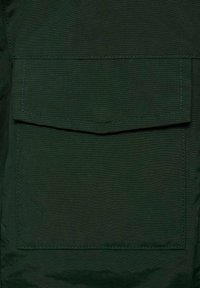Tessuto verde scuro caratterizzato da una grande tasca applicata con patta. La texture appare liscia con cuciture visibili attorno ai bordi.