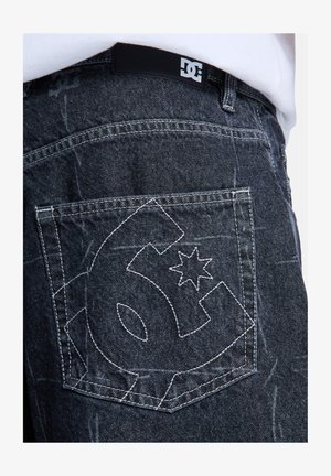 Czarna kieszeń spodni dżinsowych z białym obszytym logo DC i czarną naszywką DC na pasie, noszona z białą koszulą.