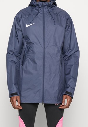 Hombre con una chaqueta con capucha con cremallera de Nike azul marino con bolsillos laterales y mallas negras con detalles rosas.
