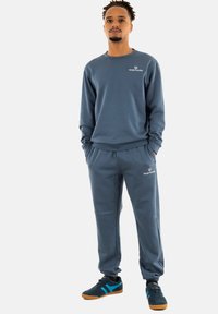 Grauer Sweatshirt und passende Jogginghosen aus weichem Material, mit Logo-Stickerei auf der Brust und dem Oberschenkel, kombiniert mit schwarzen und blauen Sneakern.