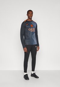 Uomo alto che indossa una maglia da calcio a maniche lunghe di colore scuro con accenti arancioni, pantaloni neri Nike, calze bianche e scarpe da ginnastica nere, in posizione eretta.