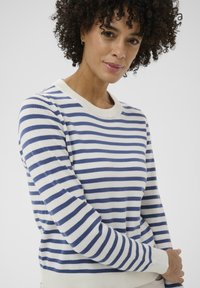 Femme aux cheveux bouclés portant un pull blanc à rayures horizontales bleues, debout devant un fond gris clair uni.