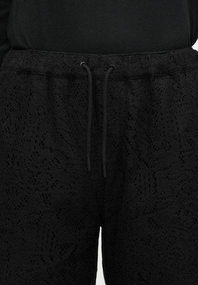 Μαύροι υφασμάτινοι παντελόνες με υφή και ελαστική μέση με κορδόνι, φορεμένοι με μαύρο τοπ, που δείχνουν λεπτομερή δαντελωτό μοτίβο υφάσματος.