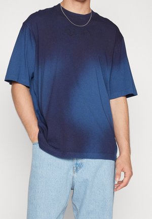 Mężczyzna w granatowo-niebieskiej koszulce z krótkim rękawem, przejście kolorów, jasnoniebieskie dżinsy oraz cienki srebrny łańcuszek, ręce opierają się po bokach.