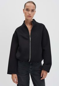 Zwarte cropped jack met een hoge kraag, ritssluiting aan de voorkant en wijde mouwen, gemaakt van textuurstof, gecombineerd met donkere jeans.