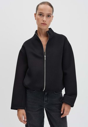 Giacca nera corta con colletto alto, zip frontale e maniche ampie, realizzata in tessuto testurizzato, abbinata a jeans scuri.