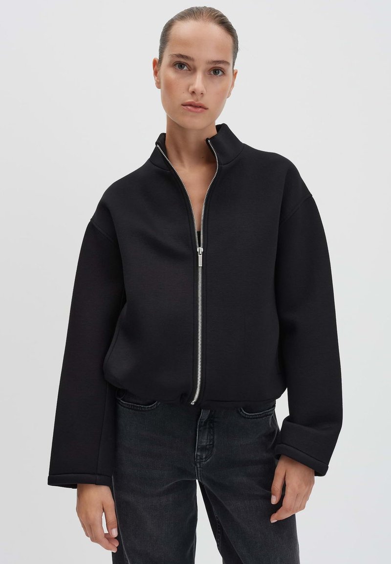 Zwarte cropped jack met een hoge kraag, ritssluiting aan de voorkant en wijde mouwen, gemaakt van textuurstof, gecombineerd met donkere jeans.