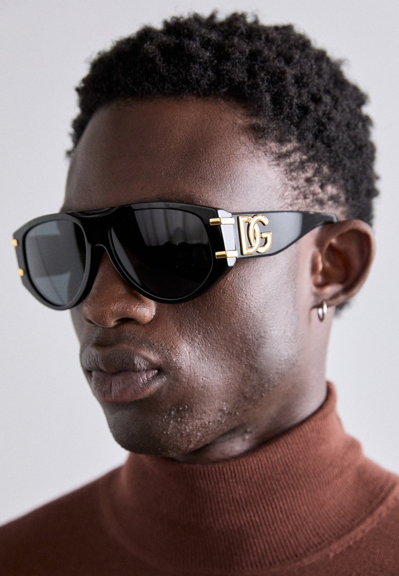 Gafas de sol negras con montura gruesa y lentes oscuros grandes. Presentan acentos de hardware dorado y un logotipo en el lateral. Acabado mate.