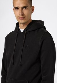 Uomo che indossa un hoodie nera di Champion con lacci, che guarda dritto di fronte a uno sfondo bianco uniforme.