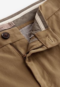Pantalon slim kaki avec fermeture à boutons et braguette partiellement zippée, montrant la ceinture intérieure et la doublure en tissu rayé.