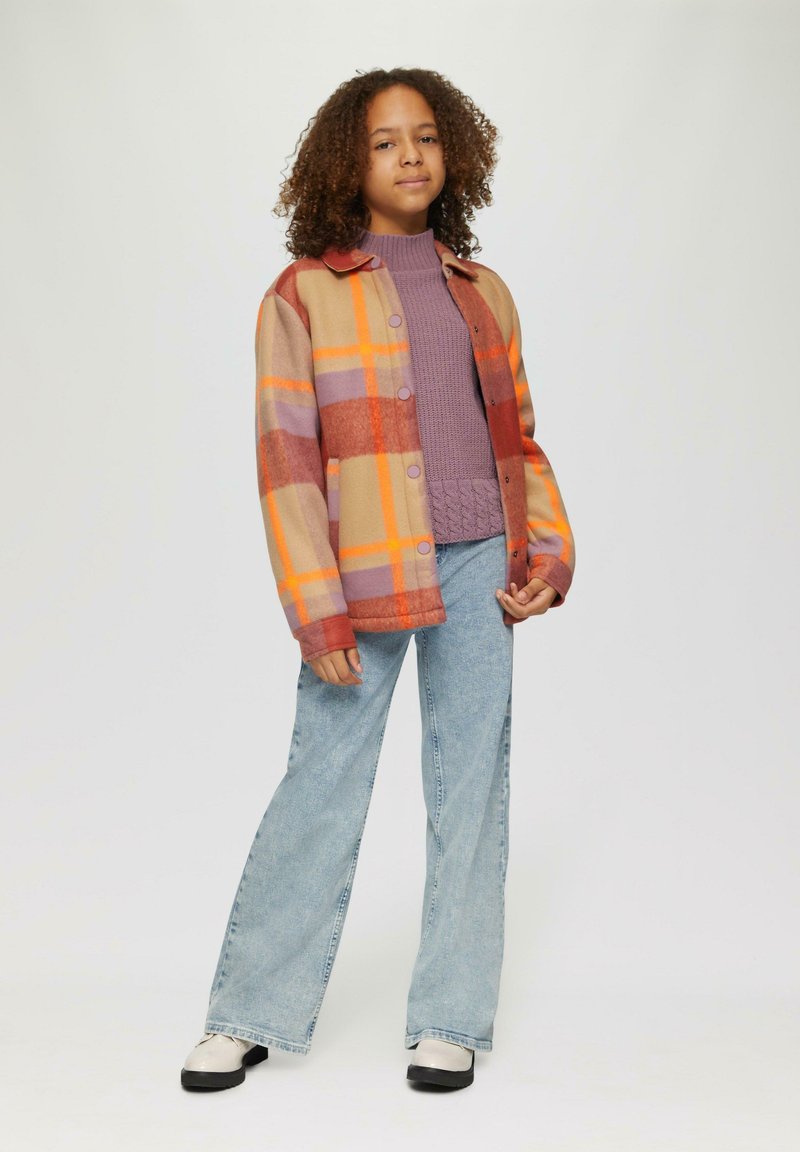 Veste en polaire à carreaux orange, violet et beige, associée à un pull en tricot lavande et à un jean léger à jambes larges bleu clair. Chaussures noires et blanches.