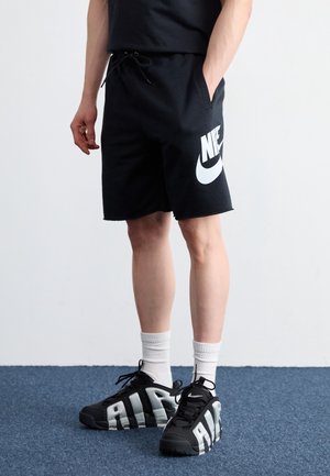 Osoba ubrana w czarne spodenki Nike z białym logo, białe skarpetki do kostek i czarne buty Nike Air, stojąca na niebieskim dywanie.