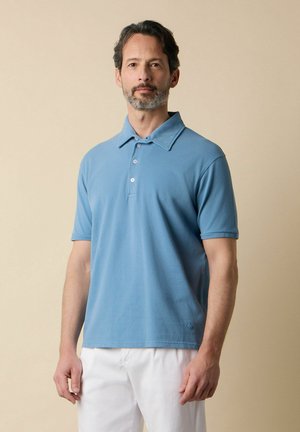 Homme aux cheveux courts et foncés avec une barbe, portant un polo bleu clair à manches courtes et un pantalon blanc, se tenant devant un fond beige.