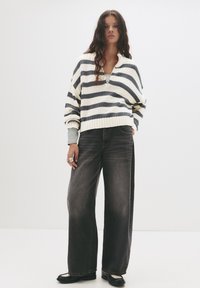 Gestreifter Strickpullover in Creme und Marineblau, V-Ausschnitt, oversized Passform. Dunkelgraue weit geschnittene Jeans mit hoher Taille. Schwarze Schuhe mit kontrastierendem Sohlen.