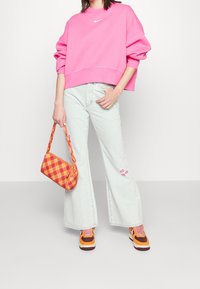 Osoba v jasně růžovém oversized svetru, světlých rozšířených džínách s vyšívanýma očima, s oranžovo-fialovými teniskami a s oranžovou kostkovanou kabelkou.