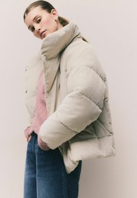 Helle beige Daunenjacke mit gerippter Textur, hohem Kragen und Knöpfen. Kombiniert mit einem rosa, flauschigen Pullover und blauen Jeans.