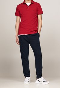 Rode poloshirt, marineblauwe chino's en witte sneakers. De poloshirt heeft een kraag en korte mouwen; de chino's zijn recht. Minimaal merkdetail.