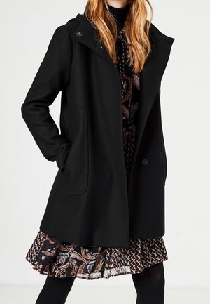 Kobieta w czarnym płaszczu z kapturem na wzorzystej sukience z czarnymi rajstopami, ręka w kieszeni płaszcza, na jednolitym białym tle.