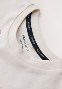 Primo piano del colletto a girocollo color crema della maglietta con etichetta nera del marchio AllSaints e la scritta "Made in Turkey" all’interno.