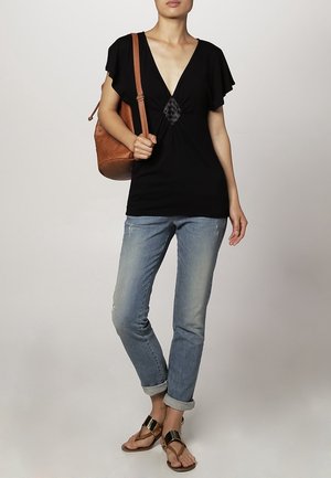 Musta V-pääntie paita, jossa on lyhyet liehuvat hihat ja keskellä poimuttaminen; yhdistetty vaaleansinisiin, kuluneisiin farkkuihin ja tasapohjaisiin sandaaleihin. Ruskea olkalaukku.